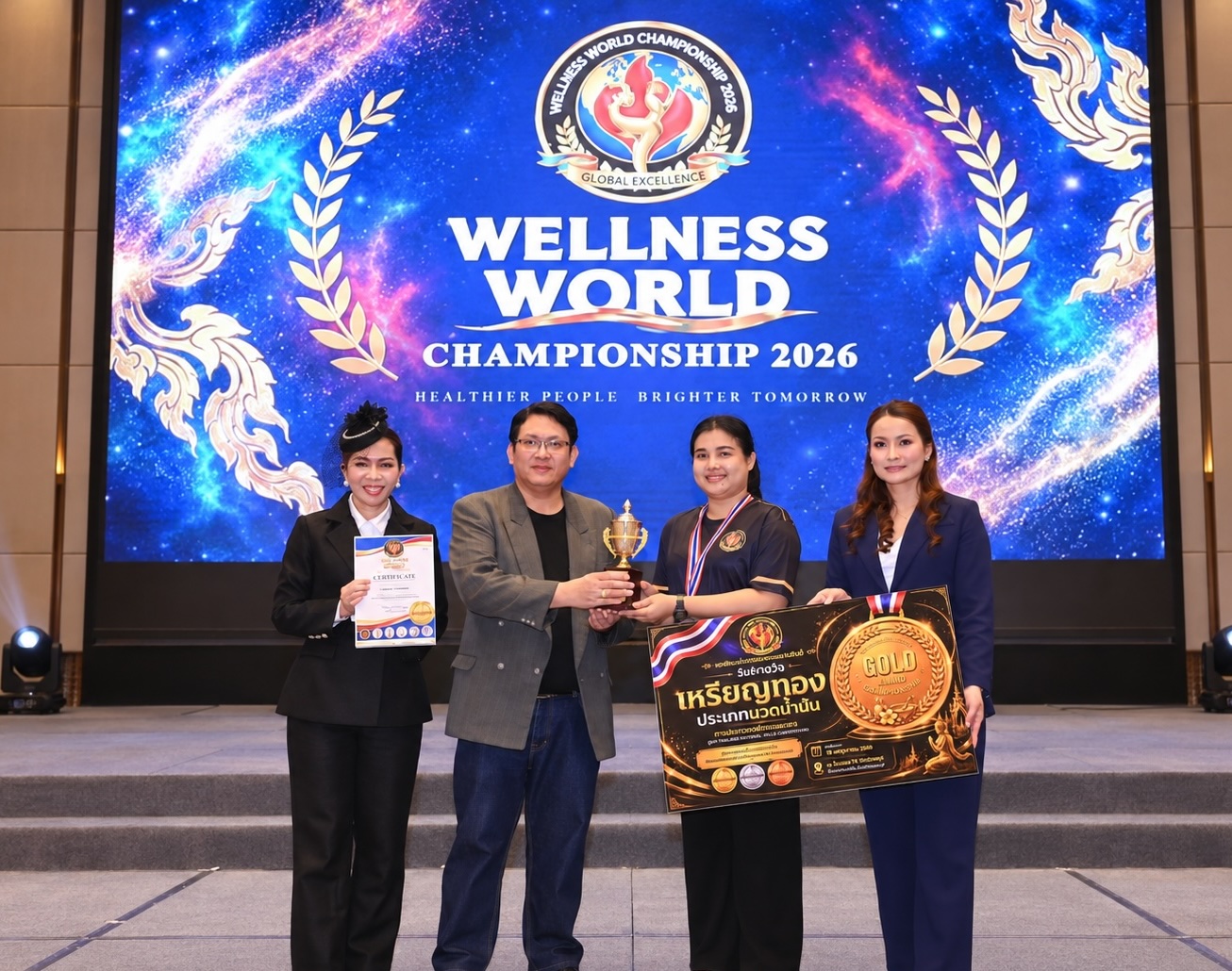 Wellness World Championship 2026 の表彰式。オイルマッサージ部門の金メダルを示すボードとトロフィーを持つ受賞者と関係者